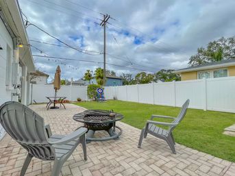 Suburban backyard with paver patio, two gray plastic patio chairs around a metal fire pit, green lawn, white vinyl fence, patio table with umbrella, target board, string lights and utility pole under a cloudy sky.