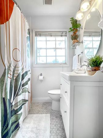 Bright modern small bathroom with tropical leaf shower curtain, white vanity and sink, hexagon tile floor, window flooding natural light, and potted plants on wall shelves.