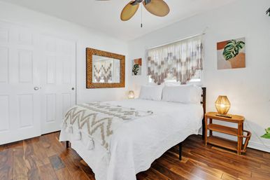 Bright boho bedroom with white bedding and tasseled throw, macramé window fringe, rattan nightstand and lamp, woven mirror, hardwood floors, palm‑leaf ceiling fan and tropical artwork