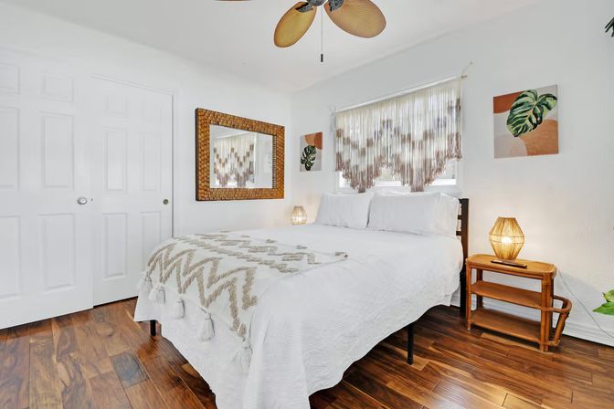 Bright boho bedroom with white bedding and tasseled throw, macramé window fringe, rattan nightstand and lamp, woven mirror, hardwood floors, palm‑leaf ceiling fan and tropical artwork