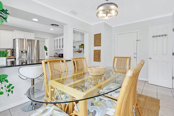 Bright coastal-style dining room with glass-top table, woven rattan chairs and wicker centerpiece, open white kitchen with stainless-steel refrigerator and breakfast bar with modern white stools.