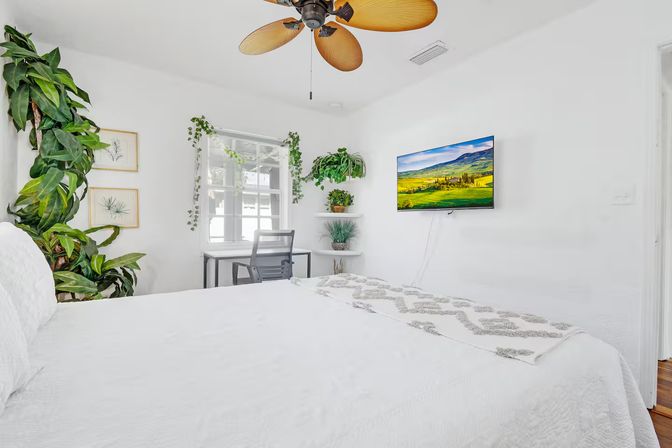 Bright, sunlit white bedroom with tropical wood-blade ceiling fan, wall-mounted TV showing a green landscape, small desk by a window, and abundant hanging and potted indoor plants.