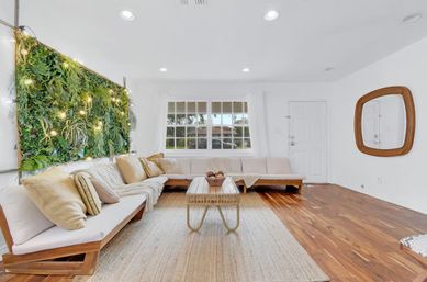 Bright, cozy modern living room with L-shaped beige sectional, wooden coffee table on a woven rug, large window, indoor vertical green wall lit with string lights, white walls and warm hardwood floors.
