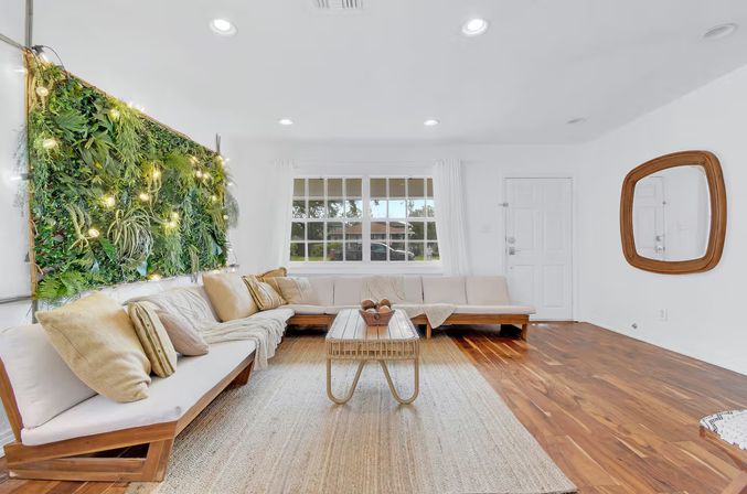 Bright, cozy modern living room with L-shaped beige sectional, wooden coffee table on a woven rug, large window, indoor vertical green wall lit with string lights, white walls and warm hardwood floors.