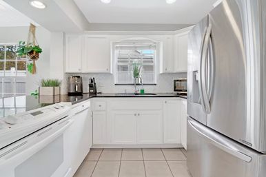 Bright modern white kitchen in a sunny home — stainless steel French-door refrigerator, black countertops, white cabinets, built-in oven and dishwasher, tile floor, window above sink and hanging plant.