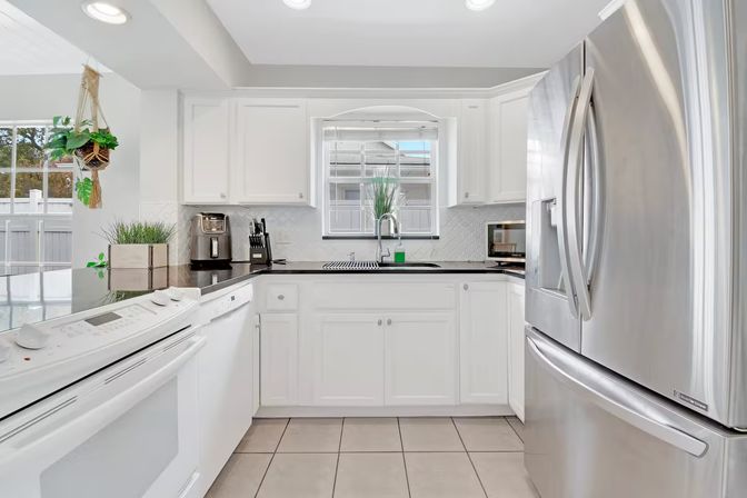 Bright modern white kitchen in a sunny home — stainless steel French-door refrigerator, black countertops, white cabinets, built-in oven and dishwasher, tile floor, window above sink and hanging plant.