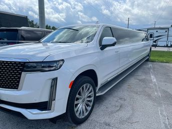 White stretched luxury SUV limousine parked in a parking lot beside RVs, long tinted windows, chrome wheels and a cloudy sky overhead