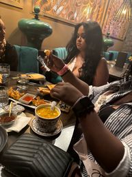 Friends dining at a modern Asian bistro — hands picking up a fried dumpling above a patterned bowl of golden soup, appetizer plates and dipping sauces on a wooden table, teal banquette and decorative green vases in the background.