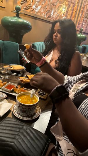 Friends dining at a modern Asian bistro — hands picking up a fried dumpling above a patterned bowl of golden soup, appetizer plates and dipping sauces on a wooden table, teal banquette and decorative green vases in the background.