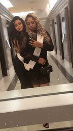 Two friends taking a mirror selfie in a hotel hallway, both in black party dresses; one with blonde curls wearing a white celebration sash and carrying a small black purse, the other with long dark hair hugging from behind — playful glam night-out vibe.