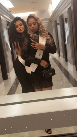 Two friends taking a mirror selfie in a hotel hallway, both in black party dresses; one with blonde curls wearing a white celebration sash and carrying a small black purse, the other with long dark hair hugging from behind — playful glam night-out vibe.