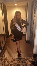 Smiling bachelorette in a black lace dress wearing a Bride Tribe sash, holding a small clutch and wearing black heels in a hotel hallway