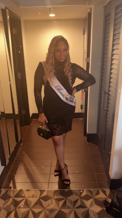 Smiling bachelorette in a black lace dress wearing a Bride Tribe sash, holding a small clutch and wearing black heels in a hotel hallway