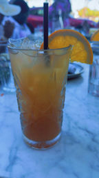 Iced orange cocktail in a tall cut-glass glass with a black straw and orange slice garnish on a marble table at an outdoor patio.