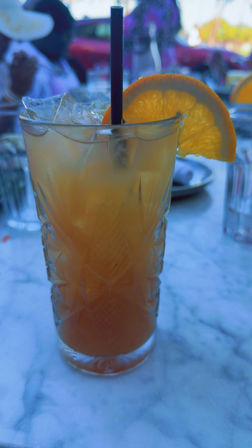 Iced orange cocktail in a tall cut-glass glass with a black straw and orange slice garnish on a marble table at an outdoor patio.