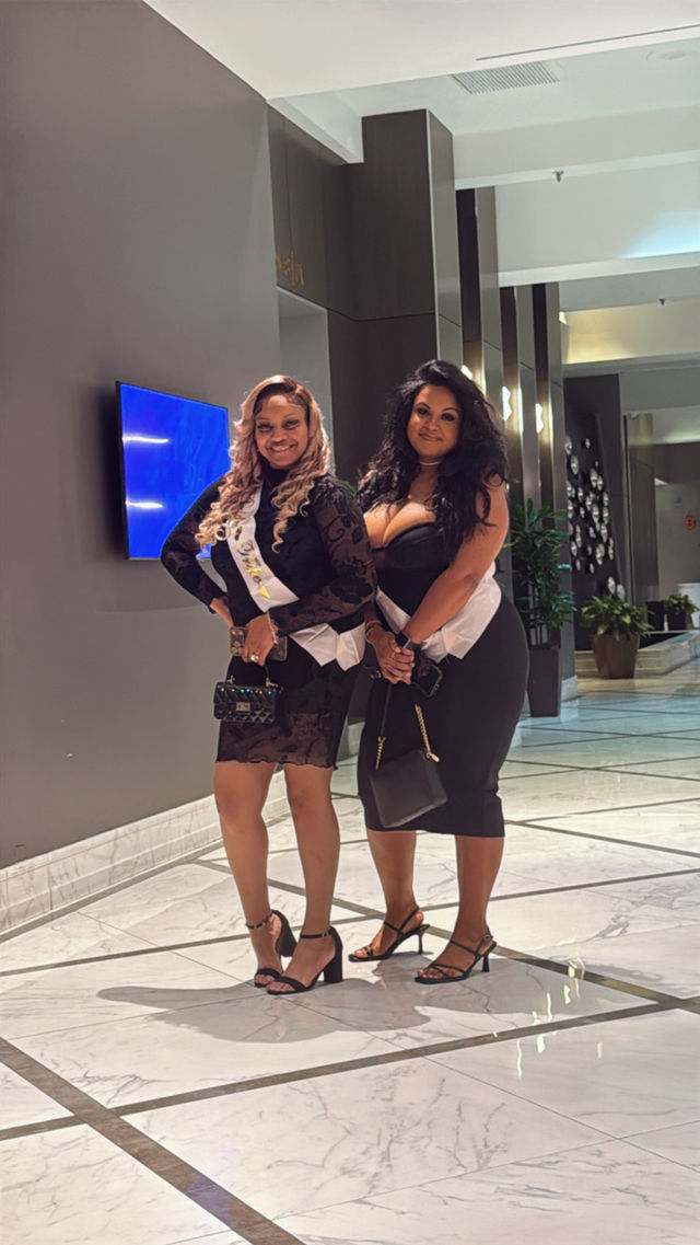 Two women in black cocktail dresses wearing white celebration sashes, heels and handbags posing in a modern hotel lobby with marble floors, potted plants and contemporary lighting
