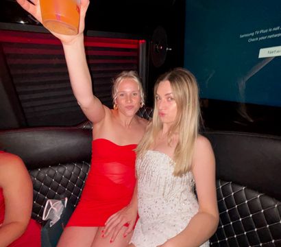 Two friends in a nightclub booth — one in a red mini dress raising a plastic cup of orange drink, the other in a white sequin mini dress on black quilted seating under red mood lighting.