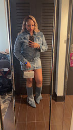 Mirror selfie of a woman in a distressed denim jacket and mini skirt, fringe denim boots and blonde waves, holding a sparkling rhinestone clutch and smartphone in a hotel room.