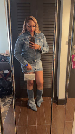 Mirror selfie of a woman in a distressed denim jacket and mini skirt, fringe denim boots and blonde waves, holding a sparkling rhinestone clutch and smartphone in a hotel room.