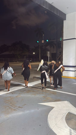 Six women wearing celebration sashes walk across a lit parking area toward a palm-lined city street at night under green traffic lights.