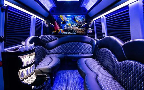 Luxury party bus interior with blue LED lighting, quilted black leather wraparound seating, illuminated mini-bar with glassware, and a colorful underwater mural at the rear.