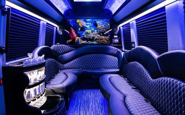 Luxury party bus interior with blue LED lighting, quilted black leather wraparound seating, illuminated mini-bar with glassware, and a colorful underwater mural at the rear.