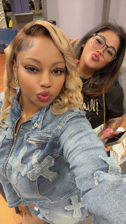 Two friends posing for a selfie in an indoor seating area — woman in front with curled blonde hair in a patched denim jacket puckering glossy lips; friend behind with dark hair, glasses, visible tattoo sleeve holding a phone and white purse.