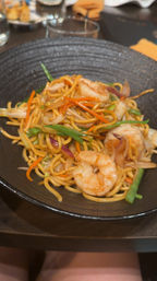 Shrimp lo mein: stir-fried egg noodles with plump shrimp, julienned carrots, green beans and onions in a glossy sauce, served in a black bowl on a restaurant table