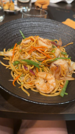 Shrimp lo mein: stir-fried egg noodles with plump shrimp, julienned carrots, green beans and onions in a glossy sauce, served in a black bowl on a restaurant table