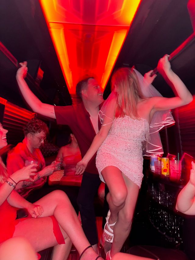 Bachelorette party on a neon-red party bus, bride in a white beaded dress and veil dancing on a seat while friends laugh and hold onto overhead rails