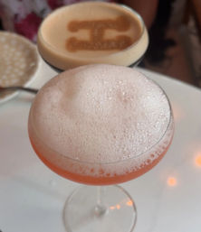 Frothy pink craft cocktail served in a coupe glass on a white cafe table, with a blurred espresso topped with cocoa-stenciled design in the background