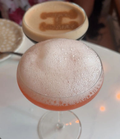 Frothy pink craft cocktail served in a coupe glass on a white cafe table, with a blurred espresso topped with cocoa-stenciled design in the background