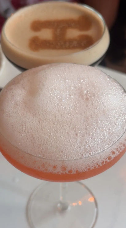 Frothy pink craft cocktail served in a coupe glass on a white cafe table, with a blurred espresso topped with cocoa-stenciled design in the background