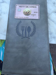 Gray leather menu cover embossed with a fork-knife-spoon wreath, topped by a pink novelty bitcoin card, resting on a white marble tabletop with a sliver of colorful patterned fabric visible.