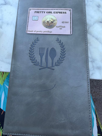 Gray leather menu cover embossed with a fork-knife-spoon wreath, topped by a pink novelty bitcoin card, resting on a white marble tabletop with a sliver of colorful patterned fabric visible.