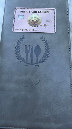 Gray leather menu cover embossed with a fork-knife-spoon wreath, topped by a pink novelty bitcoin card, resting on a white marble tabletop with a sliver of colorful patterned fabric visible.