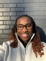 Sunlit close-up of a smiling person with glasses and braided hair in a white zip-up jacket against a dark gray urban brick wall.