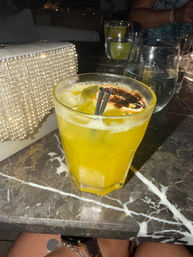 Yellow citrus cocktail in a faceted glass with ice, two black straws and a dried orange slice, on a dark marble bar table beside a sparkly rhinestone clutch and water glasses — lively night-out scene