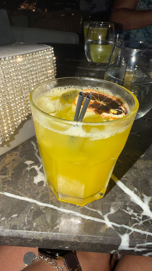 Yellow citrus cocktail in a faceted glass with ice, two black straws and a dried orange slice, on a dark marble bar table beside a sparkly rhinestone clutch and water glasses — lively night-out scene