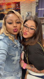 Two friends puckering for a selfie in a brightly lit indoor lobby — one in a denim jacket with blonde waves, the other wearing glasses with a tattooed arm, pink bracelets and a white quilted purse, abstract wall art behind them.