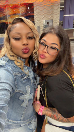 Two friends puckering for a selfie in a brightly lit indoor lobby — one in a denim jacket with blonde waves, the other wearing glasses with a tattooed arm, pink bracelets and a white quilted purse, abstract wall art behind them.