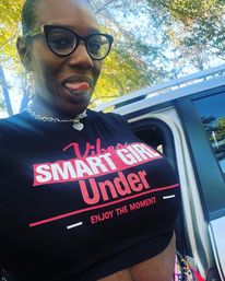 Playful outdoor selfie of a person in glasses sticking out their tongue, wearing a black crop top that reads SMART GIRL Under Enjoy the moment, standing by an open car door with sunlit trees in the background.