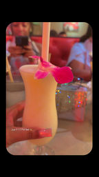 Late-night creamy tropical cocktail in a curvy hurricane glass with a straw and pink orchid garnish, held by a hand with long pink acrylic nails, blurred red booth and glittering purse in a neon-lit restaurant — text overlay 'Just 9:17 PM feel'.