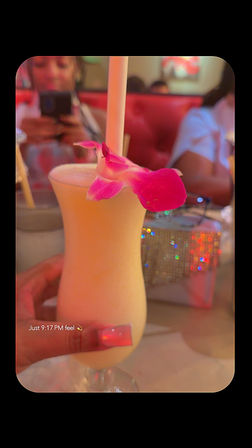 Late-night creamy tropical cocktail in a curvy hurricane glass with a straw and pink orchid garnish, held by a hand with long pink acrylic nails, blurred red booth and glittering purse in a neon-lit restaurant — text overlay 'Just 9:17 PM feel'.