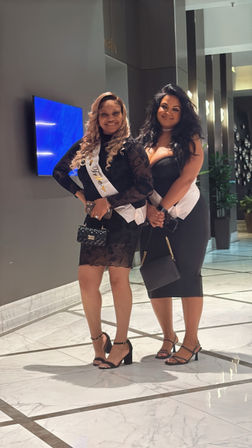 Two women in black cocktail dresses and white sashes smiling and posing in a luxury hotel lobby with marble floors and mirrored columns, holding clutch purses and wearing high heels — evening night out scene.