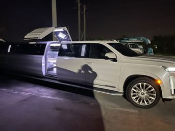 White stretch SUV limousine with gullwing roof open, revealing illuminated white leather interior and LED lighting, parked in a dimly lit lot at night with photographer's shadow on the door.