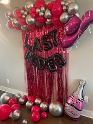 Indoor pink-and-silver party backdrop with metallic balloon garland, pink foil fringe, black letter balloons reading 'LAST RODEO', oversized pink cowboy boot and champagne bottle balloon.