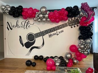 Pink, black and silver balloon arch and floor clusters framing a wall mural of an acoustic guitar with "Nashville" lettering in a living-room setting.