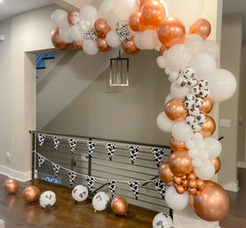 Indoor home entryway balloon garland arch in rose gold and white with cow-print balloons and matching cow-print pennant bunting draped along a modern staircase railing.