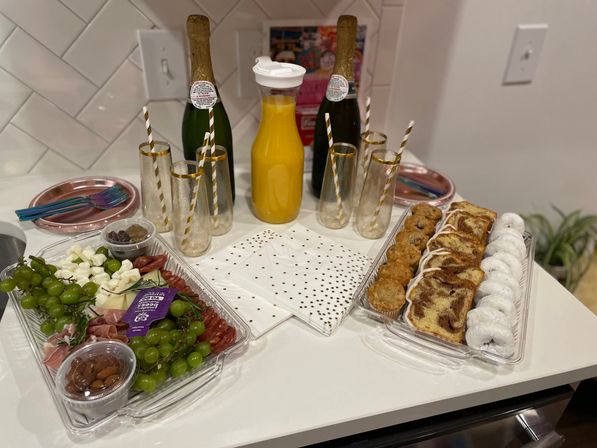 Festive brunch spread on a home kitchen island: champagne bottles, carafe of orange juice, gold-rimmed flutes with striped paper straws, charcuterie tray of grapes, cheeses, cured meats and nuts, plus a pastry platter with muffins, cinnamon swirl slices and powdered donuts.